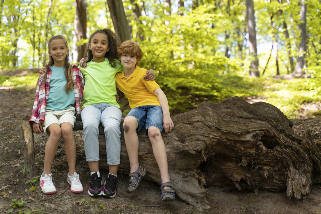 Drei fröhliche Kinder sitzen auf einem Baumstamm im Wald – Freundschaft, Abenteuer und Naturerlebnis in der Freizeit