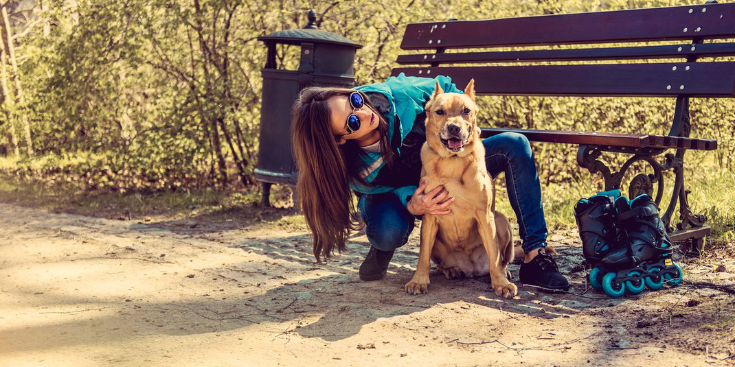 Frau mit Sonnenbrille umarmt ihren Hund neben einer Parkbank, daneben stehen Inlineskates.