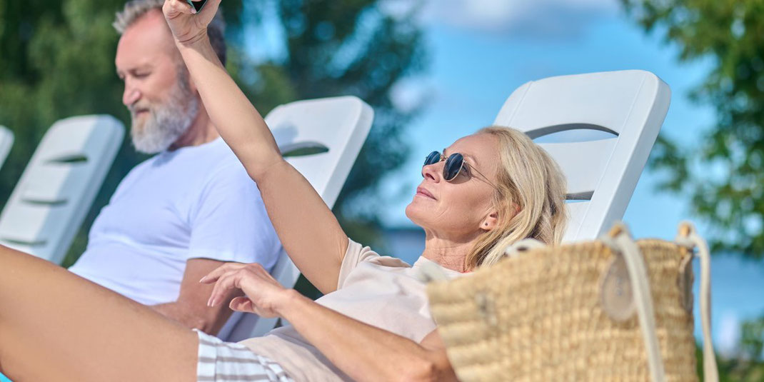 Blonde Frau in Sonnenbrille liegt entspannt auf einer Liege und macht ein Selfie.