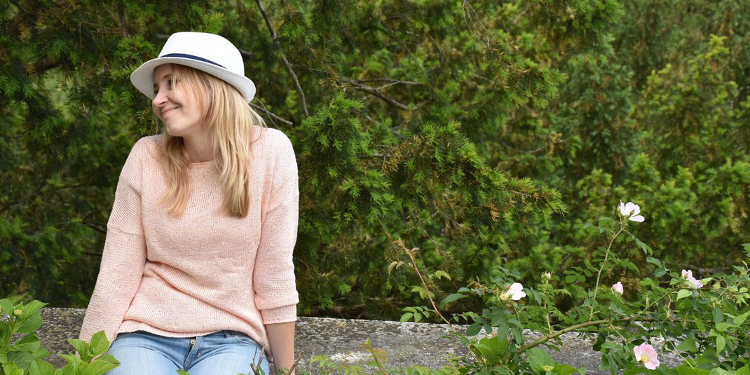 Lächelnde Frau mit weißem Hut sitzt auf einer niedrigen Mauer, umgeben von grünem Pflanzenhintergrund.