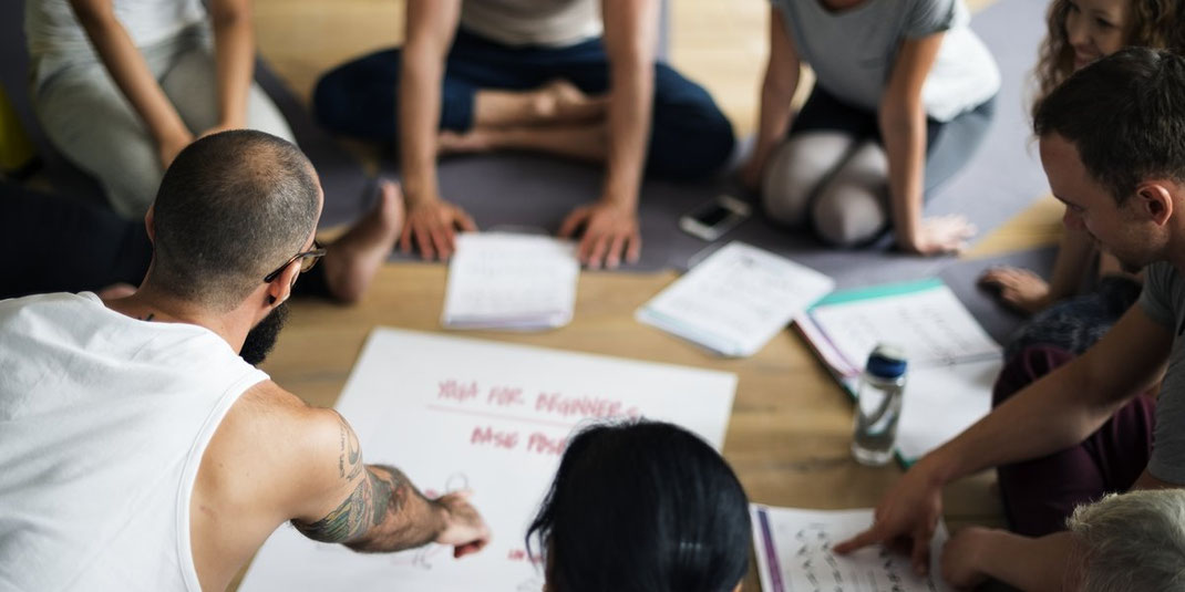 Teilnehmer einer Yoga-Klasse sitzen im Kreis auf Matten und besprechen ein Lernposter, Fokus auf Zusammenarbeit und Lernen.