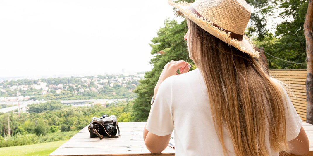 Frau mit Strohhut sitzt an Holztisch und blickt auf grüne Landschaft mit Stadt im Hintergrund.