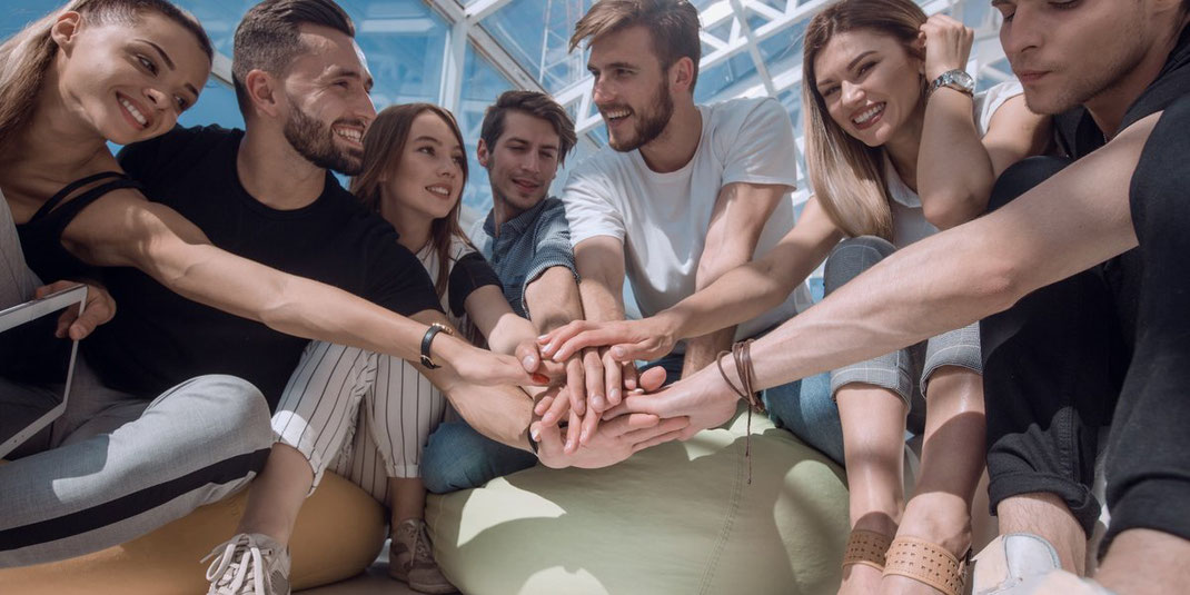 Junge Menschen sitzen im Kreis und legen motiviert die Hände in der Mitte aufeinander – Symbol für Teamwork