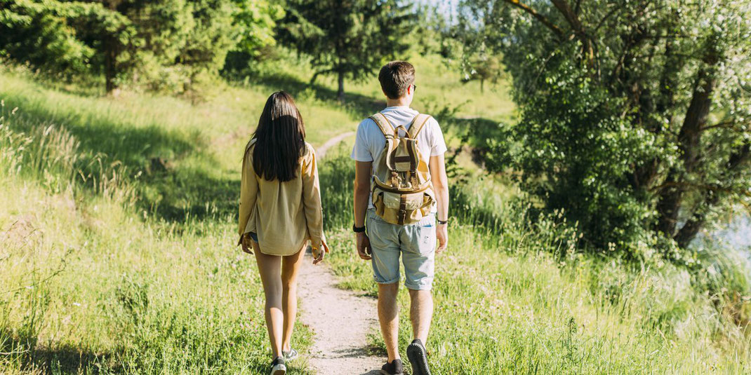 Ein Paar wandert gemeinsam auf einem schmalen Naturpfad durch eine grüne Landschaft. Die Frau trägt ein gelbes Oberteil und hat lange dunkle Haare, der Mann trägt ein weißes T-Shirt und einen Rucksack. Sie gehen zwischen hohem Gras und Bäumen entlang eine