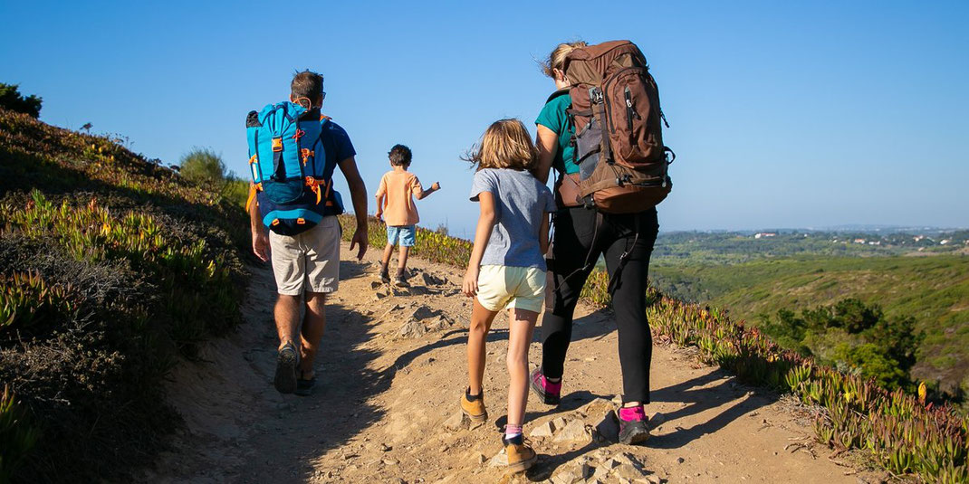 Familie mit zwei Kindern und großen Rucksäcken wandert bei Sonnenschein durch hügelige Landschaft