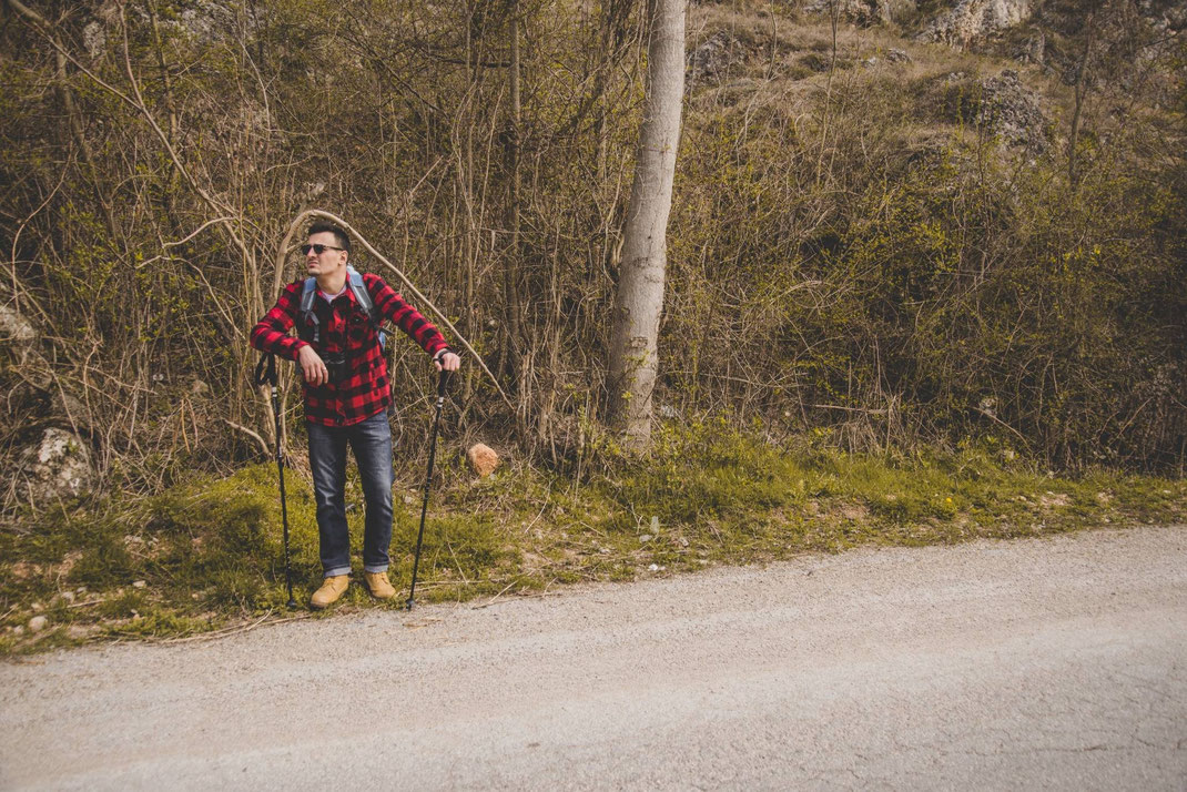 Ein Wanderer in rotem Holzfällerhemd lehnt sich mit Wanderstöcken am Wegesrand an und ruht sich in der Natur aus.