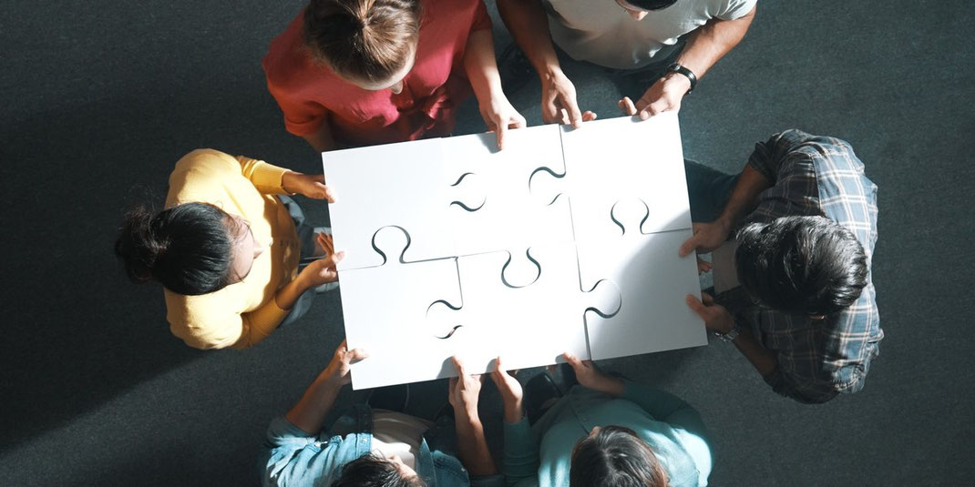 Gruppe von Menschen steht im Kreis und hält gemeinsam ein zusammengesetztes Puzzle hoch – Symbol für Zusammenarbeit