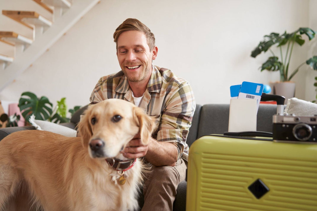 Lächelnder Mann sitzt mit seinem Golden Retriever neben einem grünen Koffer und bereitet sich auf eine Reise vor.