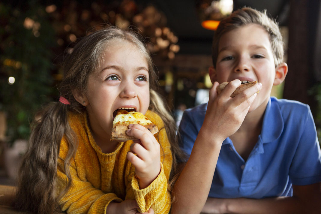 Zwei fröhliche Kinder essen Pizza in einem gemütlichen Restaurant, das Mädchen trägt einen gelben Pullover und der Junge ein blaues Poloshirt.