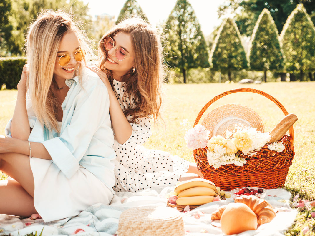 Zwei junge Frauen genießen ein sommerliches Picknick auf einer Wiese, lachen miteinander und sitzen entspannt neben einem Picknickkorb voller Obst und Blumen.