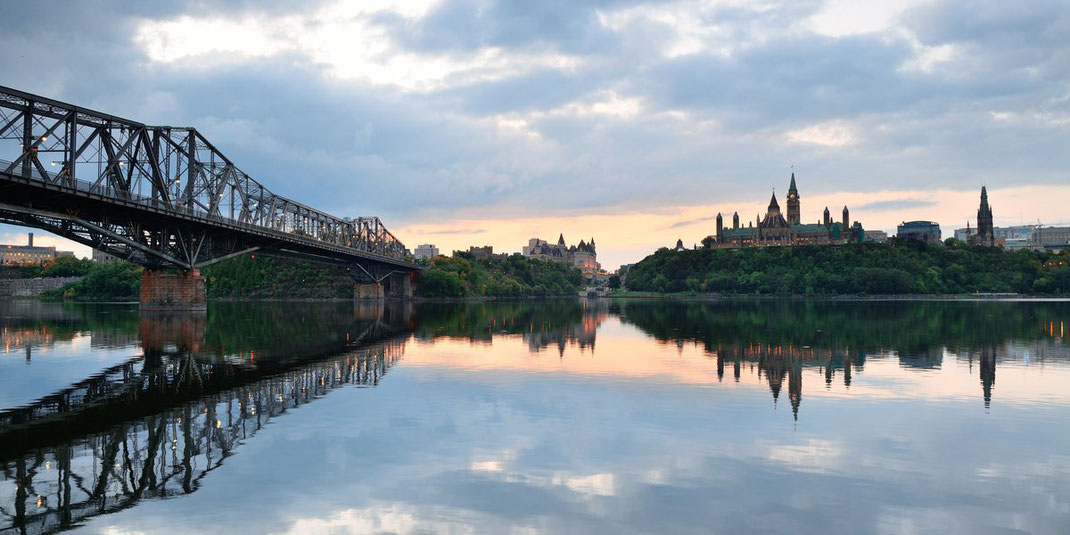 Blick auf die Skyline von Ottawa mit Parlamentsgebäuden und Brücke, Spiegelung im ruhigen Wasser bei Sonnenaufgang.