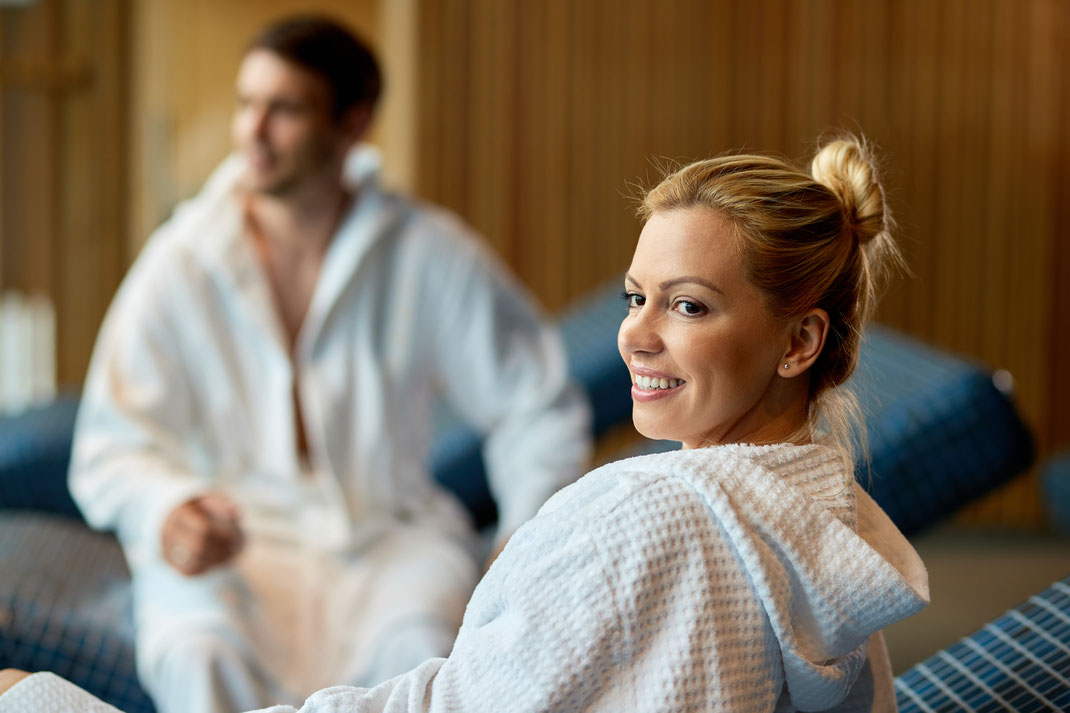 Lächelnde Frau im Bademantel mit Partner im Hintergrund in der Spa-Lounge – Paarwellness, Entspannung und Wohlfühlzeit im Wellnesshotel