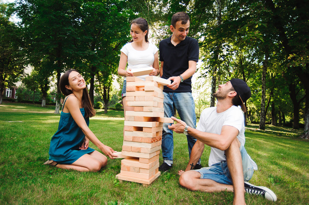 Vier junge Erwachsene spielen draußen im Park ein Riesen-Jenga-Spiel – Spaß, Geschicklichkeit und gemeinsames Lachen an einem sonnigen Tag.