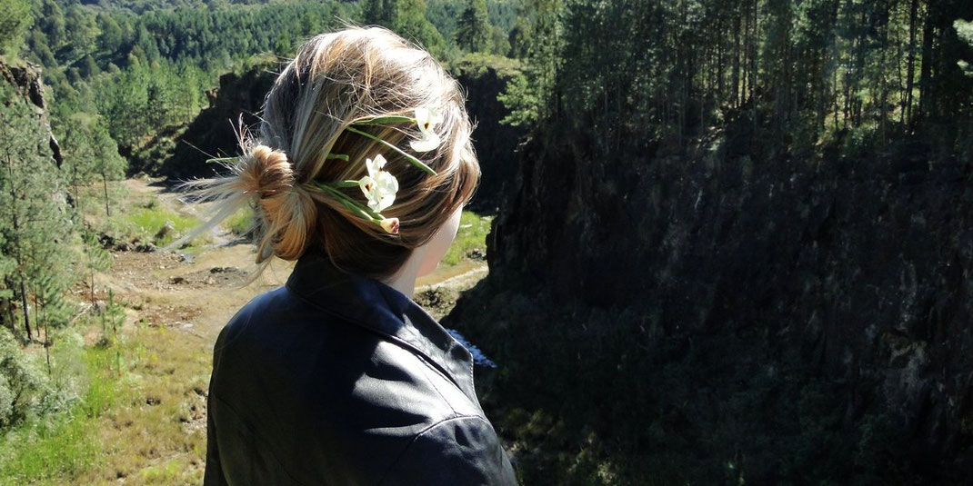Junge Frau mit Blumen im Haar steht mit dem Rücken zur Kamera und blickt auf einen bewaldeten Abhang