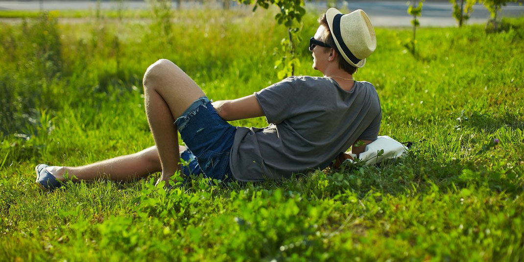 Junger Mann mit Hut und Sonnenbrille liegt entspannt auf einer grünen Wiese im Park.