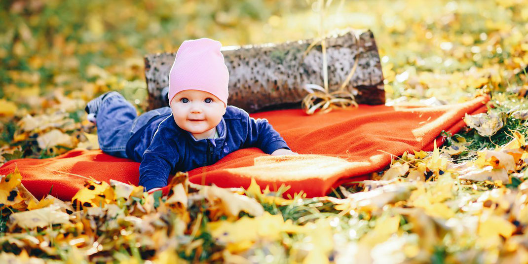 Baby mit pinker Mütze liegt auf einer roten Decke zwischen herbstlichen Blättern im Park.