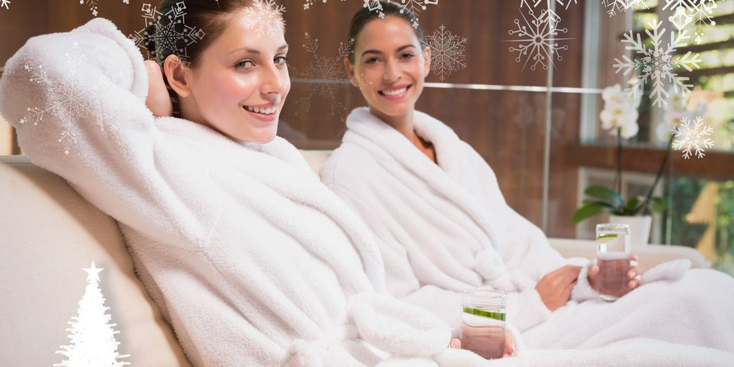 Zwei Frauen in Bademänteln sitzen lachend im Spa, dekoriert mit winterlichen Schneeflocken