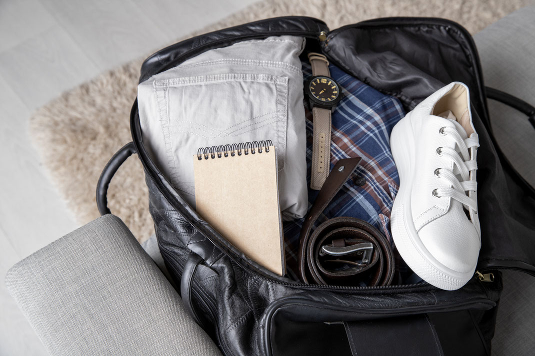 Geöffnete Reisetasche mit neatly gepackter Kleidung, Gürtel, Sneaker, Notizblock und Armbanduhr – bereit für den Wochenendtrip.