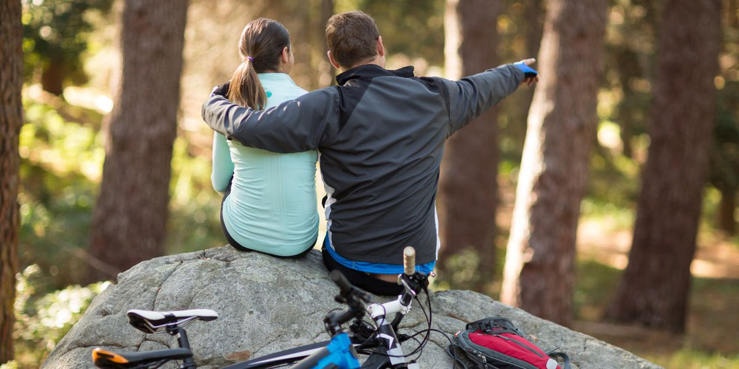 Paar mit Fahrrädern sitzt auf einem Felsen im Wald, der Mann zeigt in die Ferne