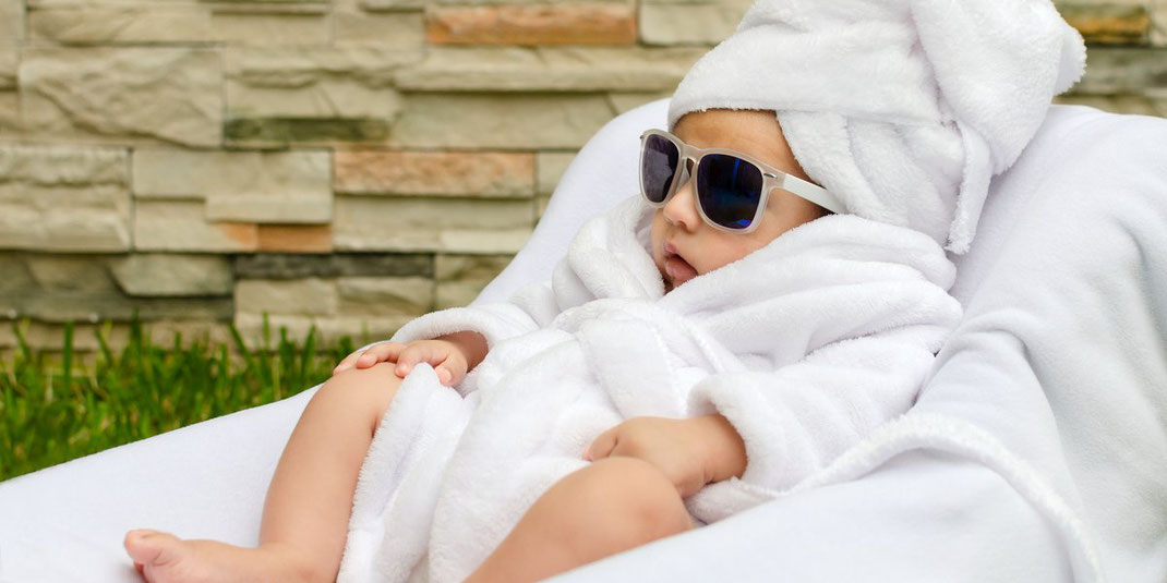 Baby in weißem Bademantel und Handtuchturban trägt Sonnenbrille und liegt entspannt auf einer Liege im Freien.
