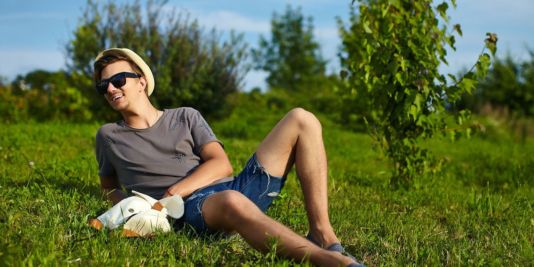 Lässiger junger Mann mit Hut und Sonnenbrille entspannt auf einer grünen Wiese im Sommer.