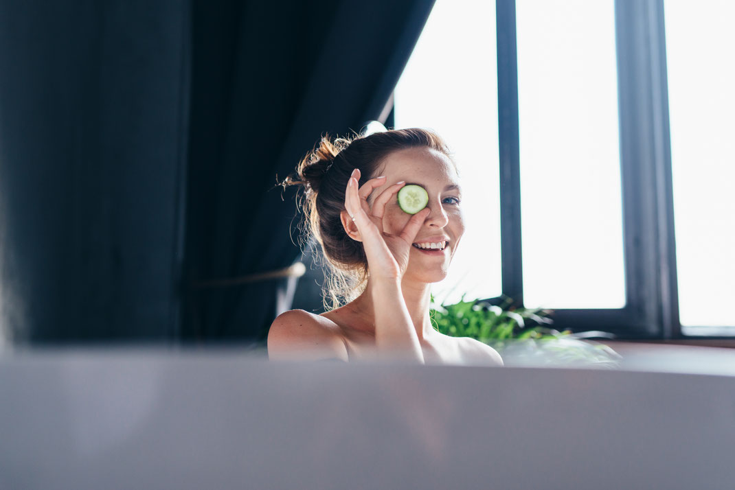 Lächelnde Frau in der Badewanne hält eine Gurkenscheibe vor ihr Auge – entspannter Moment bei einem Spa- oder Beauty-Ritual zu Hause.