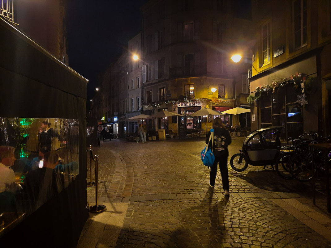 Eine junge Frau bummelt durch eine schmale Straße in einer Altstadt. Es ist Nacht. Rechts und links befinden sich Restaurants. Straßenlaternen tauchen die Szene in ein warmes, gelbliches Licht.