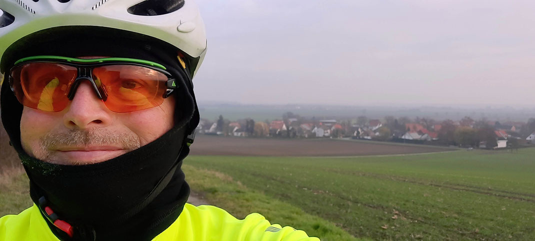 Ein Radfahrer mit Helm und Brille steht vor einem Dorf im Nebel.