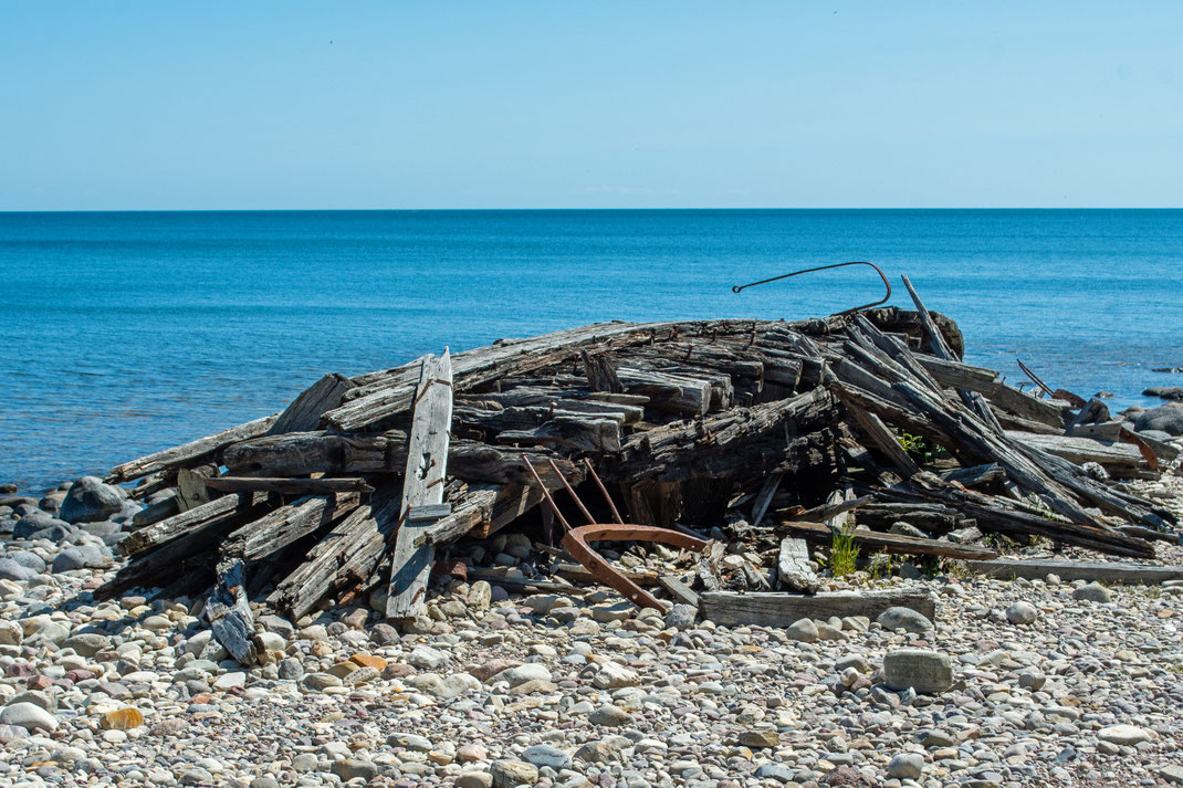 Auf einem steinigen Strand an einer Meeresküste liegt das Wrack eins Holzbootes.