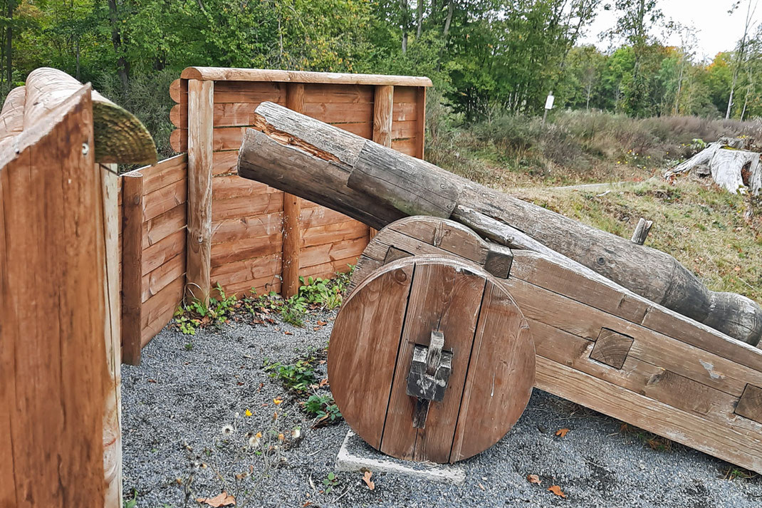 Aus Holz gefertigt ist der Nachbau eines alten Geschützes. Es steht auf einem Platz im Wald. 