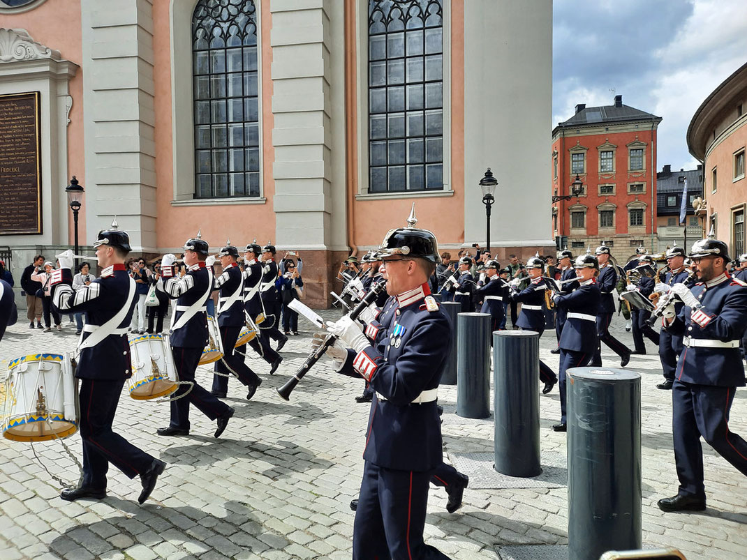 Ein Musikkorps in dunkelblauen Uniformen marschiert über einen historischen Platz mit Kopfsteinpflaster. Im Hintergrund sind eine Mauer und mehrere Fenster einer großen Kirche zu sehen.