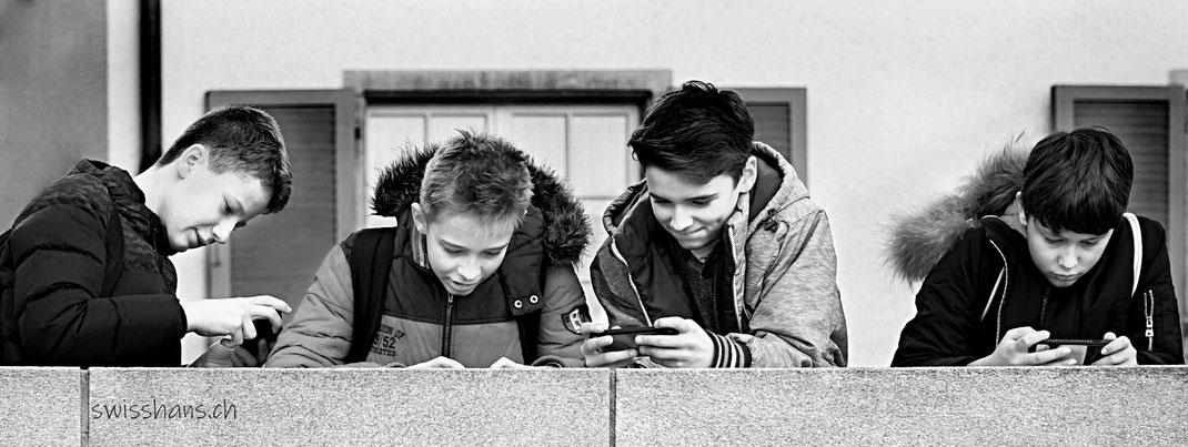 Vier Knaben mit Smartphone auf einer Mauer vor einer Hausfassade.