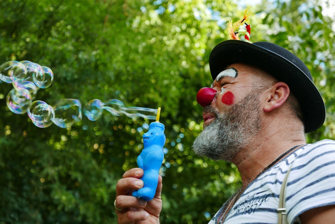 Clown mit Seifenblasen, Rimpimpim, Poesie im Park, Wiesbaden 2019. Rechts Clown mit Hut und Bart bläst mit blauem Seifenblasenbär Blasen nach links
