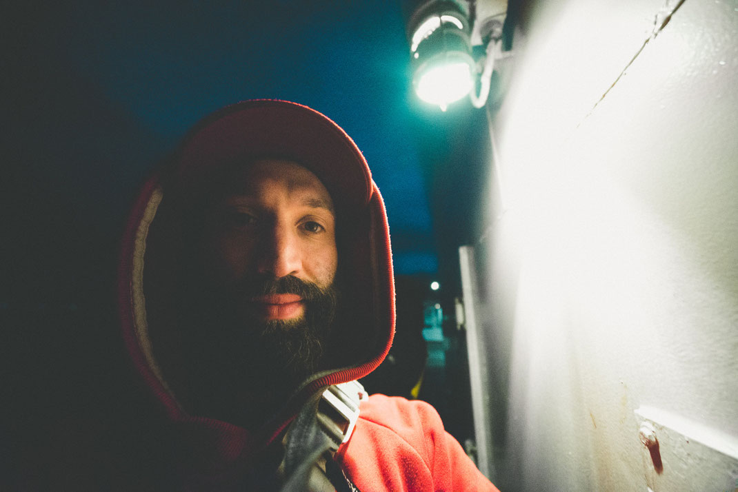a bearded man with hoodie on a ferry deck looking into the camera