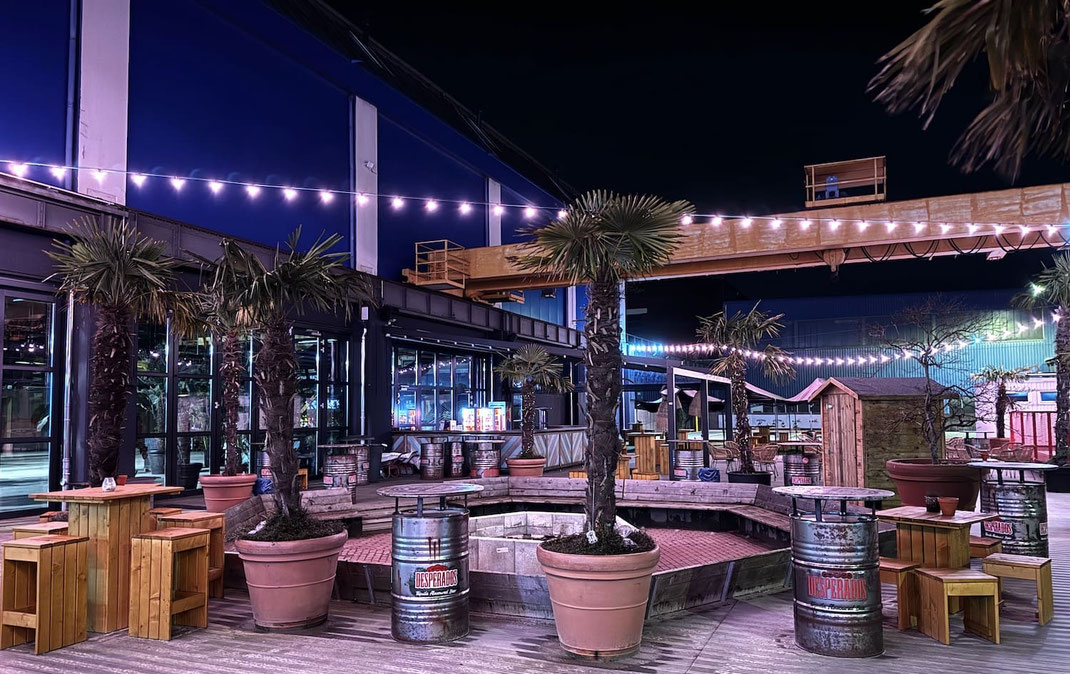 Nachtaufnahme einer Beachbar im Industriegebiet mit Palmen, Lichterketten und Stehtischen aus Ölfässern vor einer Fabrikhalle