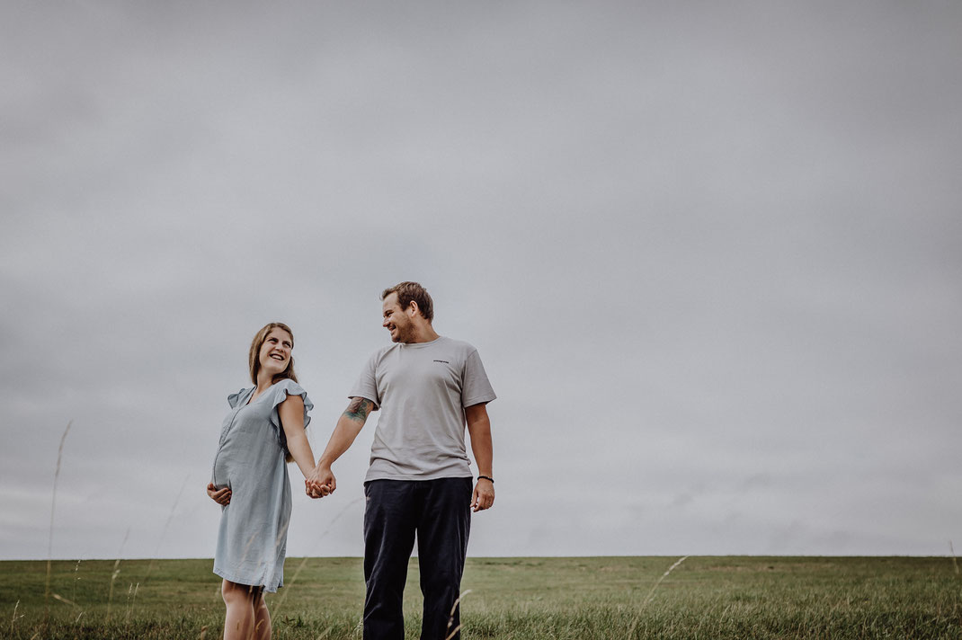 Junge werdende Eltern stehen Händchen haltend nebeneinander am Berg in Hürtgenwald und lachen sich an. Die werdende Mami hält stolz ihren Babybauch mit der rechten Hand fotografiert von Portraitfotografin Uschi Kitschke aus Kreuzau.