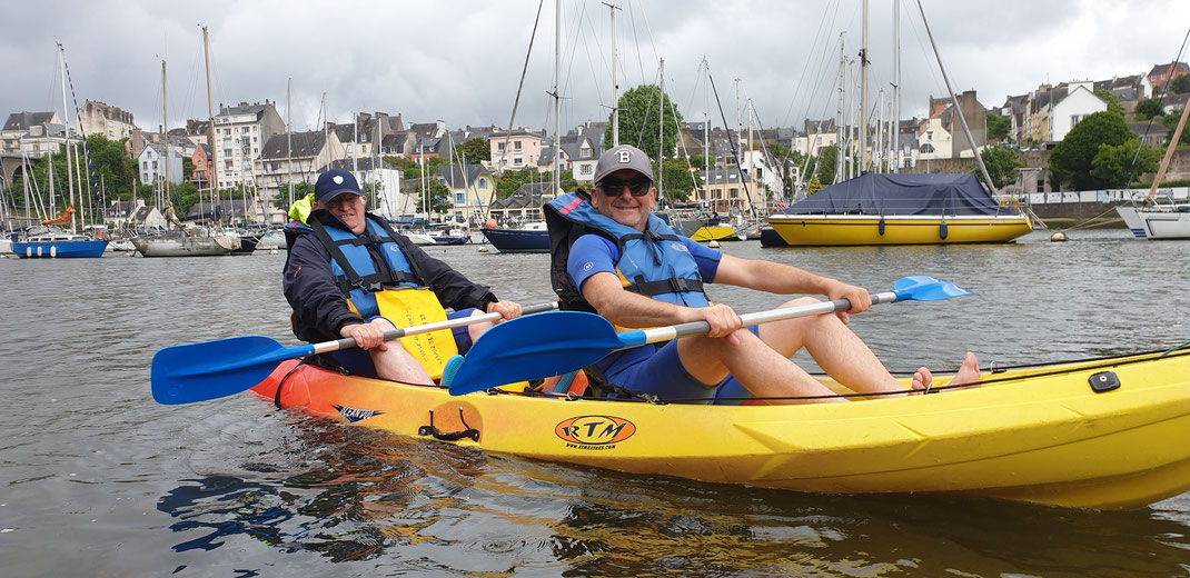 Grand kayak jaunes avec 2 hommes souriants à bord, derrière eux les maisons du Port Rhu à Douarnenez, rive Est.