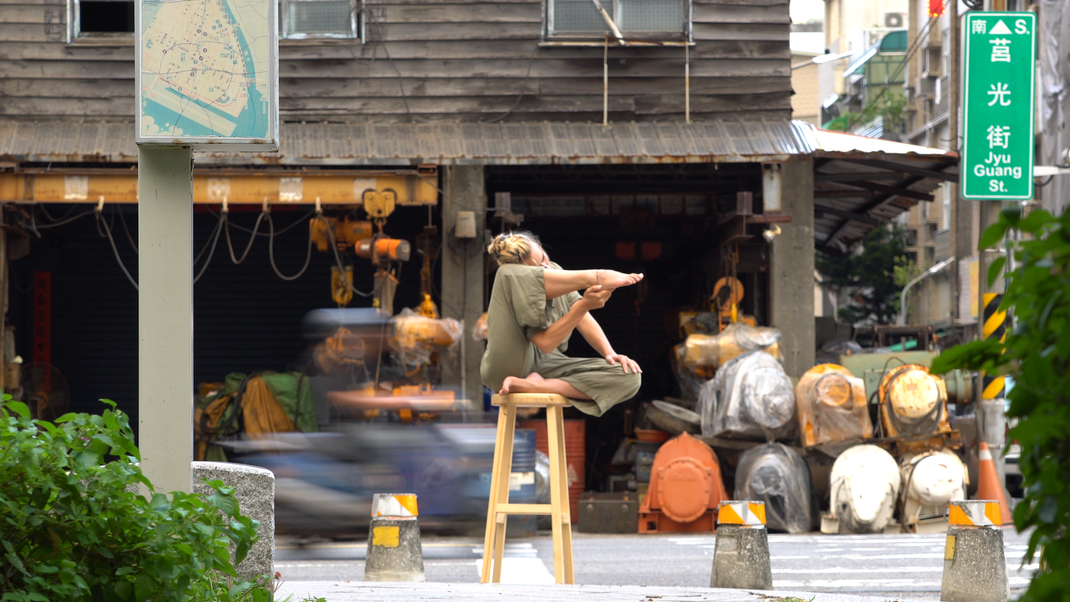 Performance Künstlerin in grünem Bodysuit sitzt in Kaohsiung City, Taiwan auf einem Barhocker, das Bein ähnlich einer Waffe aufgerichtet. Hinter ihr stehen industiell aussehende Stromgeneratoren.