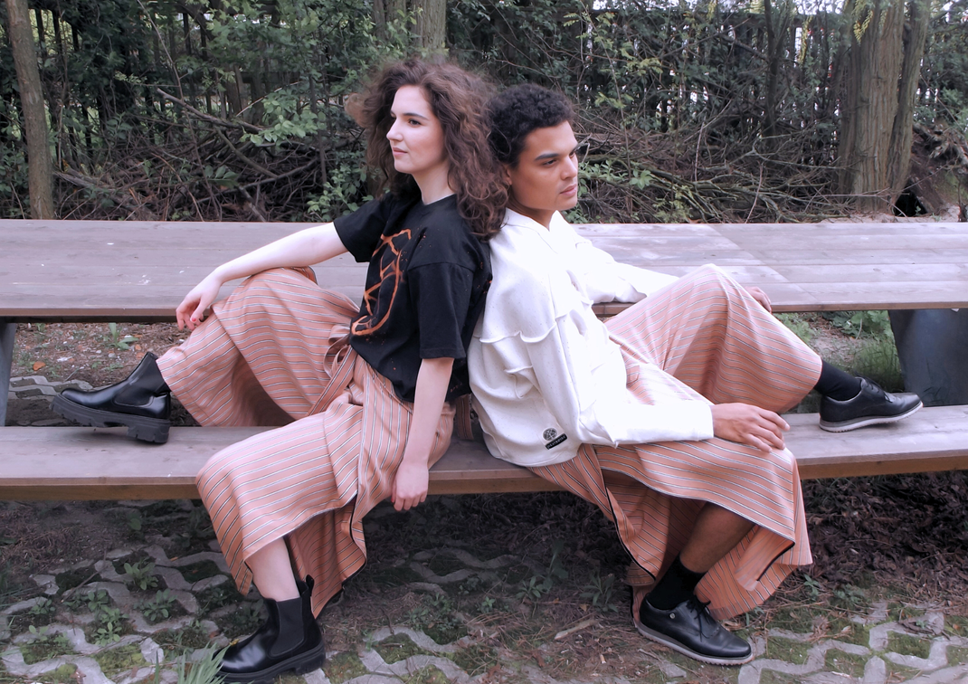 Eine Frau und ein Mann sitzen Rücken an Rücken auf einer Bank. Die Frau trägt ein schwarzes Shirt und die Sienna Pants in peach. Der Man trägt den cremefarbenen Silty Sweater und die Sienna Pants ebenso in peach.