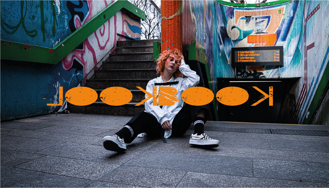 Rothaarige Frau, sitzt auf  Treppe, trägt eine schwarze Hose und ein weißes Hemd mit Ledergürteln.