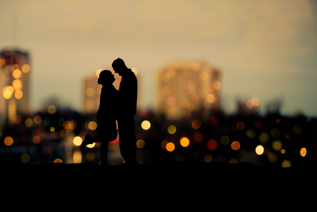 Male and female couple in silhouette in front of a bokeh city view. Buildings have an orange glow. Sky is blue. Couple is facing each other. Female has one leg kicked up behind her. 