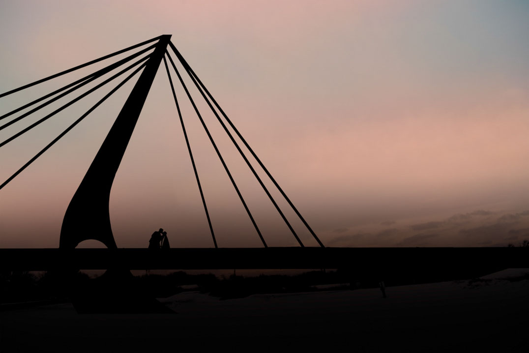 Bride and groom doing a dip kiss on a bridge at sunset. Couple is in silhouette. Bride is wearing a veil. Sky is pink and blue.