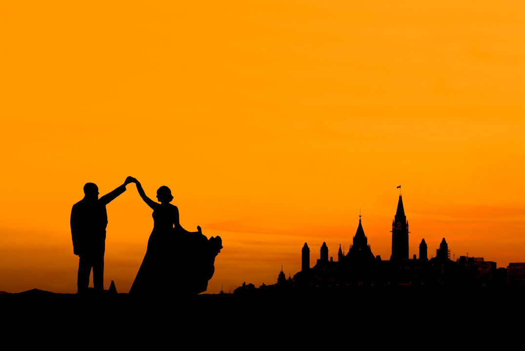 Photo of bride and groom in silhouette in front of Ottawa Parliament. Parliament hill is mostly in shadow. Sky is orange. Bride is wearing a dress and groom is wearing a suit. They are dancing. 