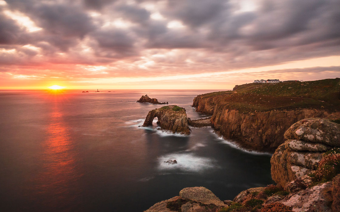 Der Enys Dodnan Arch bei Sonnenuntergang in Cornwall in England. Im Hintergrund ist der Leuchtturm Longships und Land's End zu sehen. 