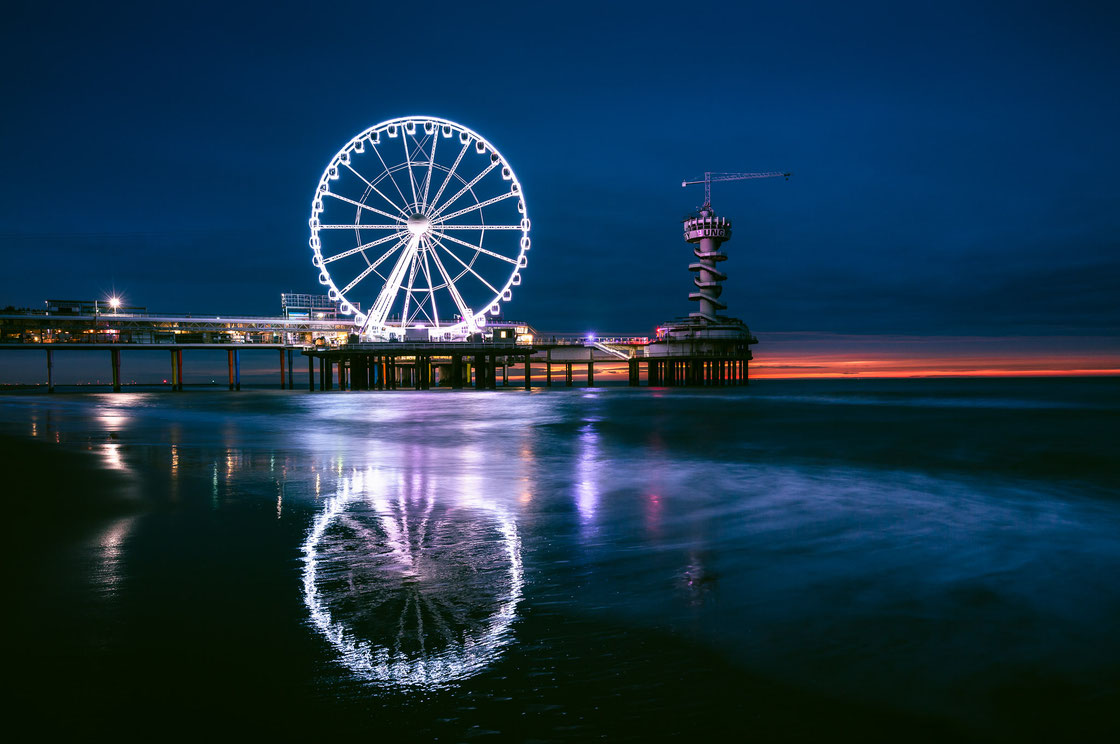 Die Seebrücke Scheveningen Pier in Den Haag in den Niederlanden in der blauen Stunde mit beleuchtetem Riesenrad und Spiegelung am Strand