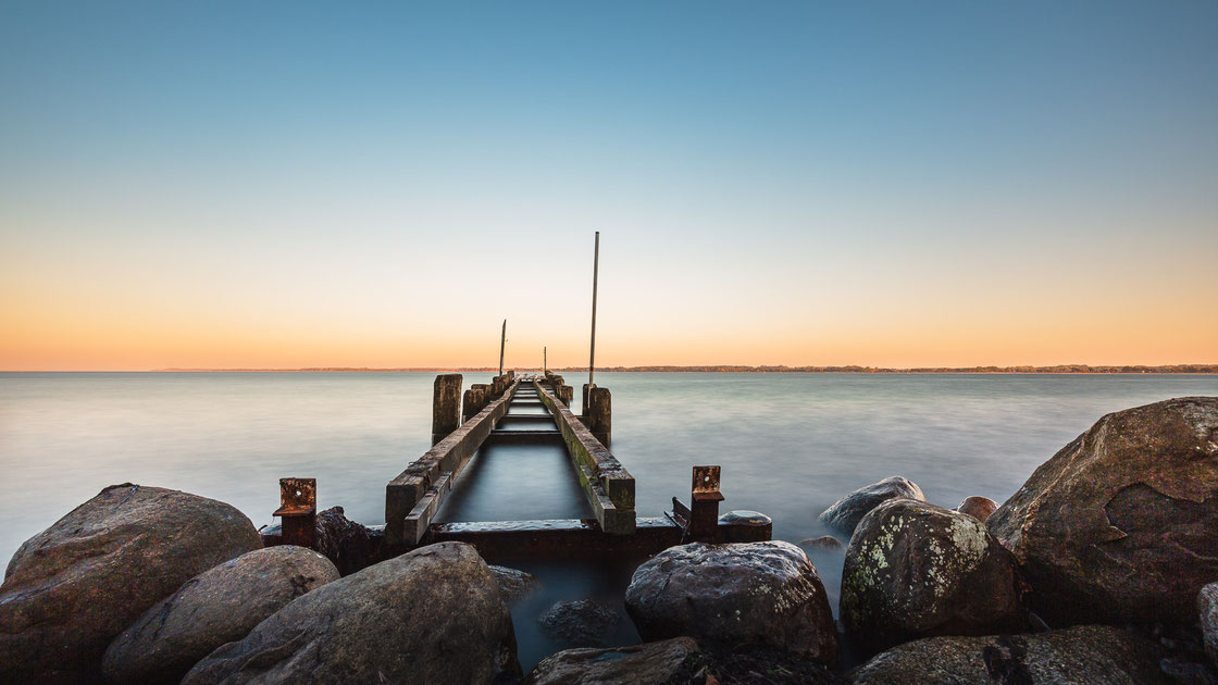 Langzeitbelichtung eines alten ehemaligen Stegs in Travemünde bei Sonnenuntergang mit Felsen im Vordergrund