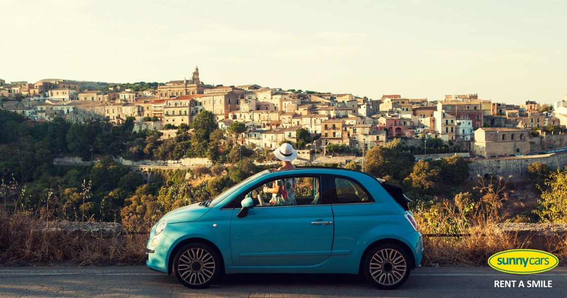 moderner blauer Mini mit einer Frau in buntem Sommerkleid und mit weißem Sonnenhut, die aufrecht im Wagen steht und aus dem Dachfenster in Richtung einer mediterranen Dorfs schaut / Bild: Sunny Cars