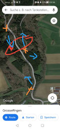 Satellitenansicht einer Landstraße mit Feldern und Wald. Mehrere rote und blaue Markierungen und Pfeile überlagern die Karte.