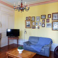 Grand salon de caractère aux murs jaunes avec canapé, poêle, écran plat et un mur décoré de portraits de famille anciens.