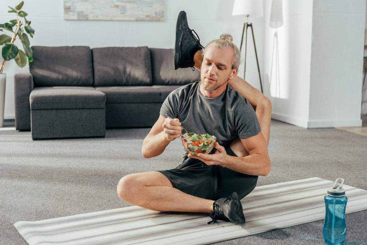 Junger Mann sitzt in sehr beweglicher Yoga Stellung auf dem Fußboden und isst dabei einen Salat